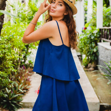 Attractive young woman in blue dress and straw hat wearing pink sunglassses walking at tropical spa villa hotel on vacation in summer style outfit, sexy