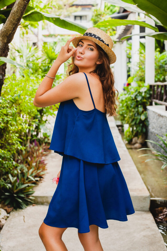 Attractive young woman in blue dress and straw hat wearing pink sunglassses walking at tropical spa villa hotel on vacation in summer style outfit, sexy